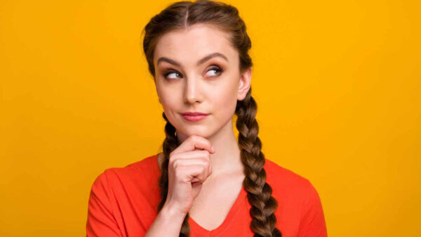 Foto de uma mulher com duas longas tranças, olhando para o lado, em um espaço vazio, com a mão no queixo, vestindo uma camiseta vermelha casual, isolada em fundo amarelo.