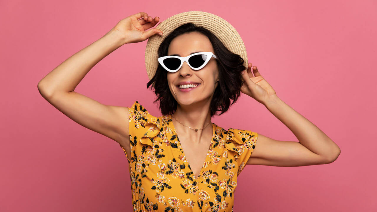 Foto de close-up de mulher muito feliz em um vestido floral, chapéu de palha e óculos escuros, que está sorrindo e tocando seu chapéu.