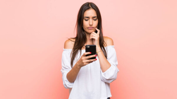Jovem mulher sobre fundo rosa, isolada, pensando e segurando celular.
