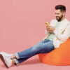 homem sentando num puff mexendo no celular em fundo rosa - Foto: Shutterstock