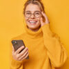 Mulher positiva sorrindo alegremente, mantendo a mão na borda dos óculos, sentindo-se alegre, usando um suéter confortável, estando contra um fundo amarelo.