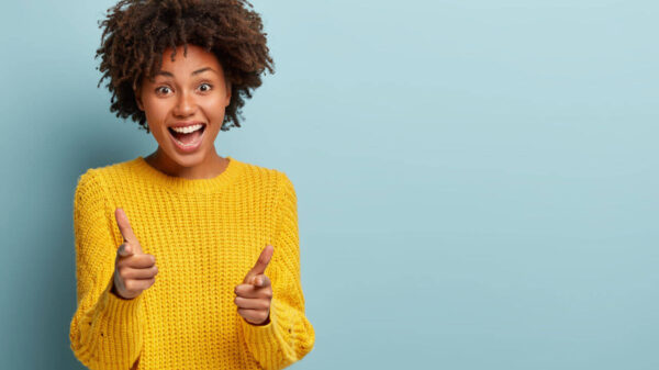 Mulher extrovertida e despreocupada fazendo pistolas de dedo, aponta diretamente para a câmera com expressão positiva, usando roupa casual, sobre a parede azul.