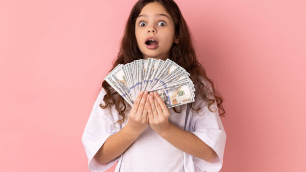 Retrato de menina rica, chocada, vestindo camiseta branca, segurando notas de dólar, olhando para a câmera com expressão de surpresa. Foto de estúdio interno isolado em fundo rosa.