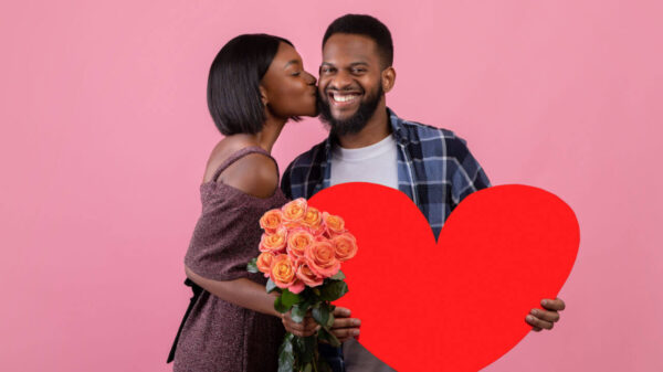 Homem segurando coração vermelho, e mulher com buquê de rosas beijando-o no fundo do estúdio rosa.