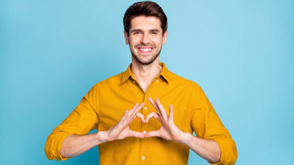 Foto de um homem sorridente fazendo um sinal em forma de coração, isolado sobre fundo de cor pastel azul.