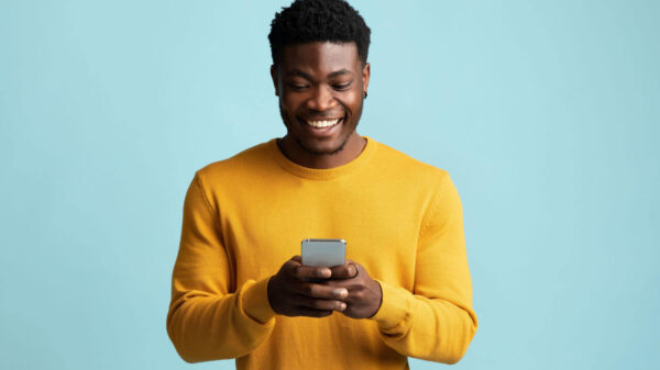 Homem segurando smartphone moderno, olhando para a tela do celular, sorrindo, em um fundo azul de um estúdio.