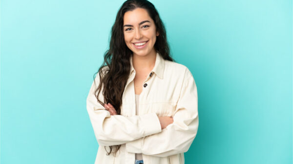 Mulher sorrindo, com os braços cruzados, sobre fundo azul claro.