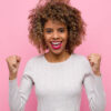 Jovem se sentindo feliz, surpresa e orgulhosa, gritando e comemorando o sucesso com um grande sorriso, contra uma parede rosa.