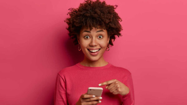 Mulher positiva, apontando para smartphone, feliz, usando blusa rosa, impressionada com algo.
