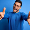 Jovem com barba, vestindo suéter casual e usando óculos, sobre fundo azul, aprovando algo, fazendo gesto positivo com as mãos, com os polegares para cima, sorrindo e feliz.