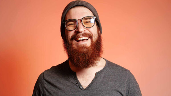 Retrato de homem feliz, barbudo, sorridente, de óculos, olhando confiante para a câmera.