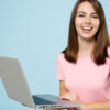Mulher sorrindo, segurando um notebook, usando blusa rosa, sobre fundo azul claro.