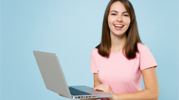 Mulher sorrindo, segurando um notebook, usando blusa rosa, sobre fundo azul claro.