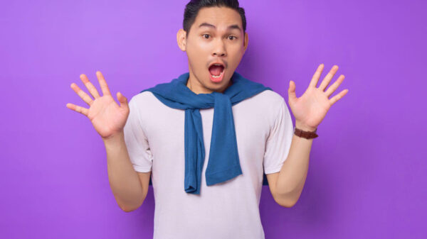 Homem jovem surpreso, vestindo uma camiseta branca, com as mãos espalhadas, isolado sobre um fundo roxo.