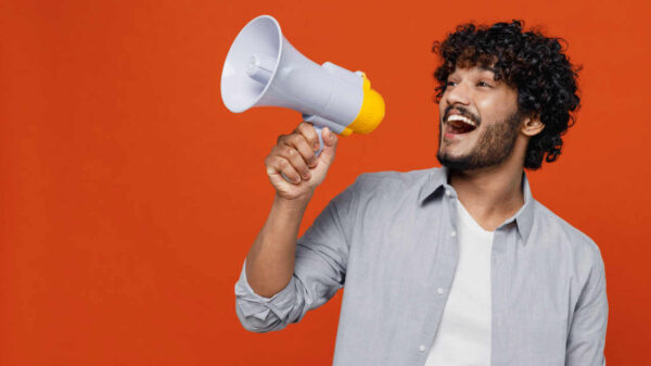 Jovem muito feliz, animado, com barba, de vestindo camisa azul, gritando em um megafone, anunciando algo, isolado em fundo vermelho/laranja. Retrato de estúdio.