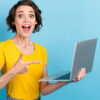 Mulher surpresa, feliz, apontando para notebook que está em sua mão, com uma blusa amarela, sobre fundo azul.