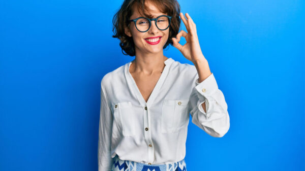 Jovem morena vestindo roupas casuais e usando óculos, sorrindo, fazendo sinal de "ok" com a mão e os dedos. Expressão de sucesso.