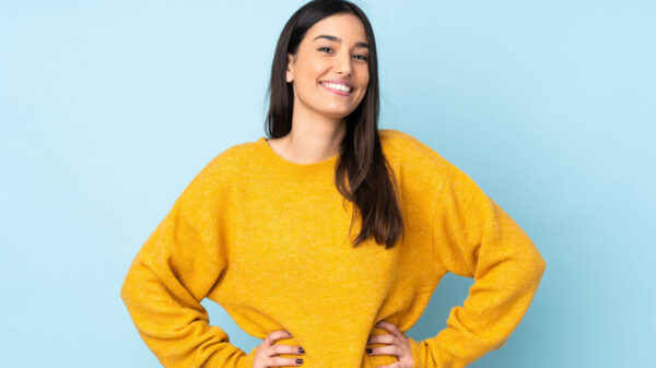 Jovem mulher isolada em fundo azul, posando com os braços no quadril e sorrindo.