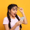 Retrato de mulher surpresa, animada, segurando e usando Smartphone, navegando nas mídias sociais. Moça emocional tirando os óculos, em fundo amarelo de um estúdio.