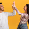 Retrato de um jovem casal feliz, fazendo "high five", alegres, em pé sobre um fundo amarelo de um estúdio.