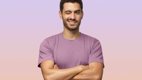 Jovem de camiseta roxa, com braços cruzados, sorrindo e piscando um olho, olhando para câmera, isolado em fundo lilás e rosa.