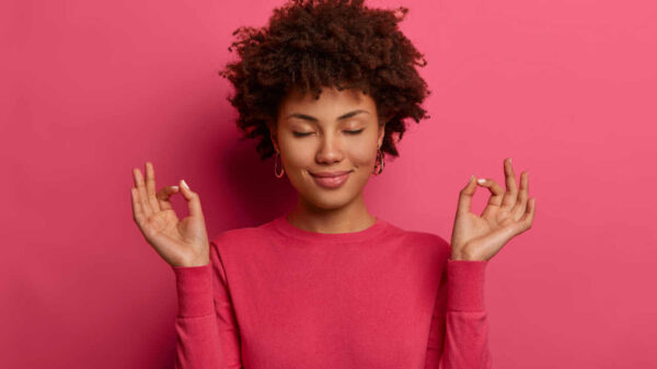 Mulher paciente, relaxada e satisfeita fazendo gesto de ioga, com os olhos fechados, usando suéter casual rosa.