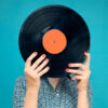 Imagem de mulher segurando disco de vinil na frente do rosto, no fundo azul.