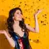 Foto de uma garota alegre, de confetes em formato de estrela voando. Garota usando uma roupa brilhante, isolada sobre fundo de cor amarela.