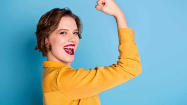 Foto de uma mulher muito alegre, mostrando o bíceps. Mulher forte e poderosa, usando camisa amarela, isolada em um fundo azul.