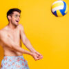 Foto de um jovem usando óculos, jogando vôlei, sorrindo, isolado em um fundo de cor amarela.
