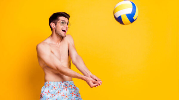 Foto de um jovem usando óculos, jogando vôlei, sorrindo, isolado em um fundo de cor amarela.