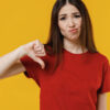 Mulher insatisfeita morena, usando camiseta vermelha básica, mostrando o polegar para baixo em gesto de negatividade, isolada em um estúdio de fundo amarelo.