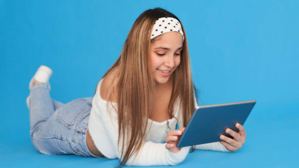 Menina mexendo no tablet, deitada no chão. Isolada no fundo azul.