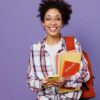 Mulher jovem, estudante, com mochila nas costas, segurando livros, usando celular, isolada em fundo roxo liso.