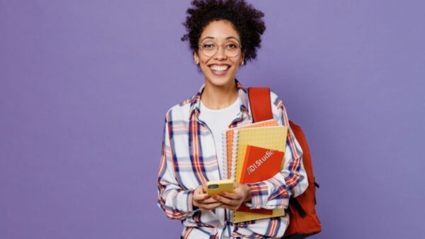 Mulher jovem, estudante, com mochila nas costas, segurando livros, usando celular, isolada em fundo roxo liso.
