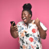 mulher negra celebrando com o celular na mão em fundo rosa