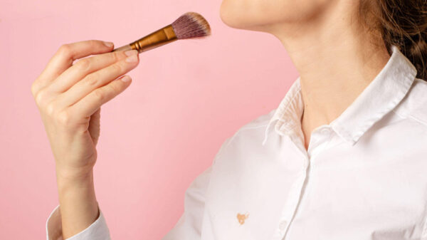 Mulher aplicando pó no rosto com pincel cosmético, isolada em fundo rosa. Usando uma camisa branca.