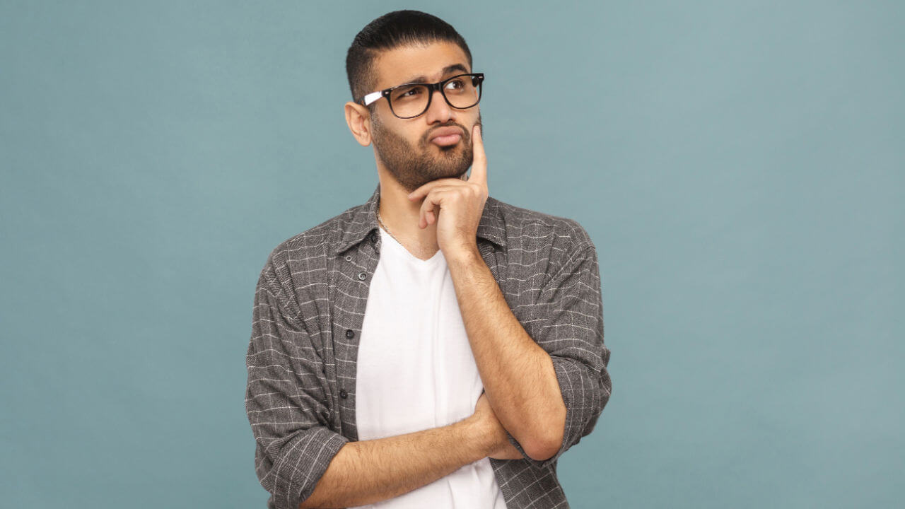 Retrato de homem pensativo, barbudo, com óculos pretos, em estilo casual, pensando. Estúdio com fundo azul.