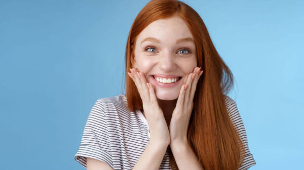 Mulher ruiva animada, feliz alegre, sorrindo com alegria, tocando as bochechas, em um fundo azul.