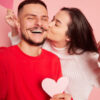 Retrato de um casal jovem. Mulher beijando homem feliz e sorridente tirando selfie dos dois juntos. Isolados sobre fundo rosa. Conceito de amor, relacionamento, emoções e estilo de vida.