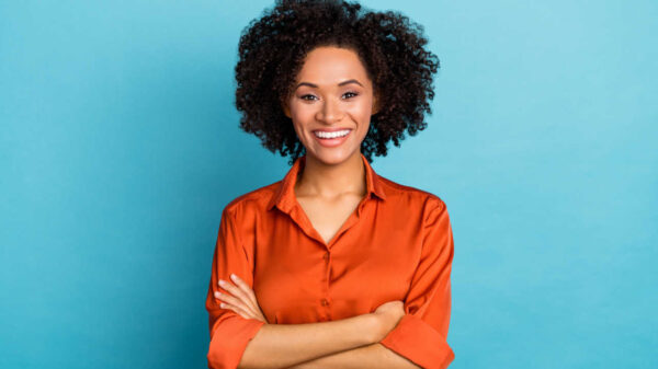 Retrato de uma mulher com os braços cruzados, sorrindo com os dentes, olhando para a câmera, isolada em fundo de cor azul.
