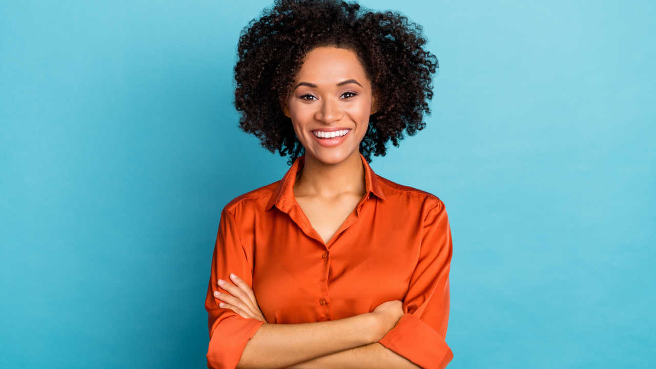 Retrato de uma mulher com os braços cruzados, sorrindo com os dentes, olhando para a câmera, isolada em fundo de cor azul.