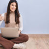 Jovem morena usando laptop, sentada no chão, fazendo gesto de polegar para cima com a mão. Expressão de aprovação. Olhando para a câmera, mostrando sucesso.