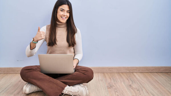 Jovem morena usando laptop, sentada no chão, fazendo gesto de polegar para cima com a mão. Expressão de aprovação. Olhando para a câmera, mostrando sucesso.