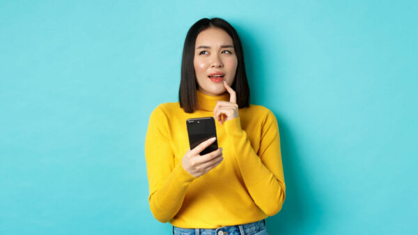 Menina pensativa, segurando smartphone, em pé sobre fundo azul.