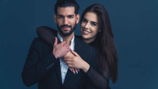Retrato de casal elegante. Mulher de cabelos compridos na casa dos 20 anos, abraçando o marido barbudo por trás. Estúdio isolado em fundo escuro.