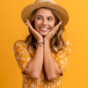 Mulher usando um vestido amarelo e um chapéu de palha, posando em um fundo amarelo isolado, com uma expressão de rosto sorridente e feliz.