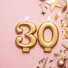 Velas de celebração douradas em formatos de número 3 e 0, e enfeites, em um fundo rosa.
