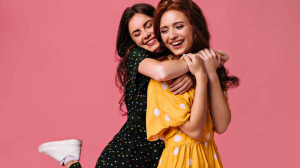 Mulheres sorrindo e se abraçando em um fundo rosa. Namoradas usando vestidos, isoladas.
