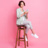 Foto em tamanho real de uma jovem sentada em um banco, usando o celular, com uma roupa cinza, isolada em um fundo rosa.